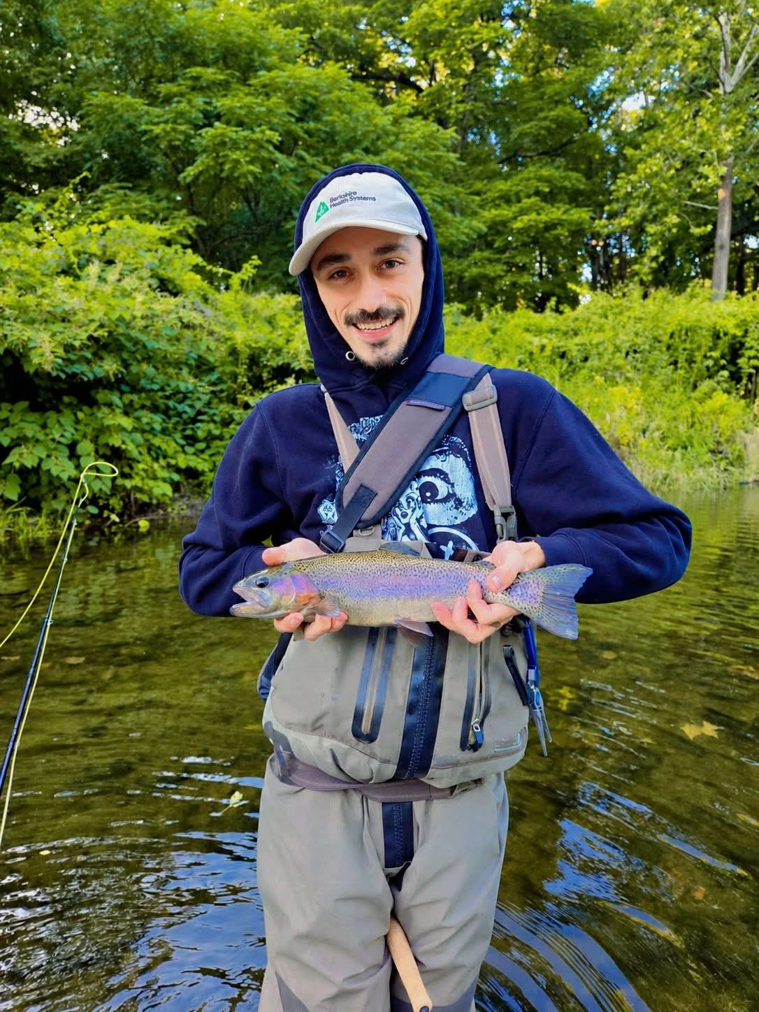 Northwater guide Nicholas Martin holding a rainbow trout while wading in a green, tree-lined river